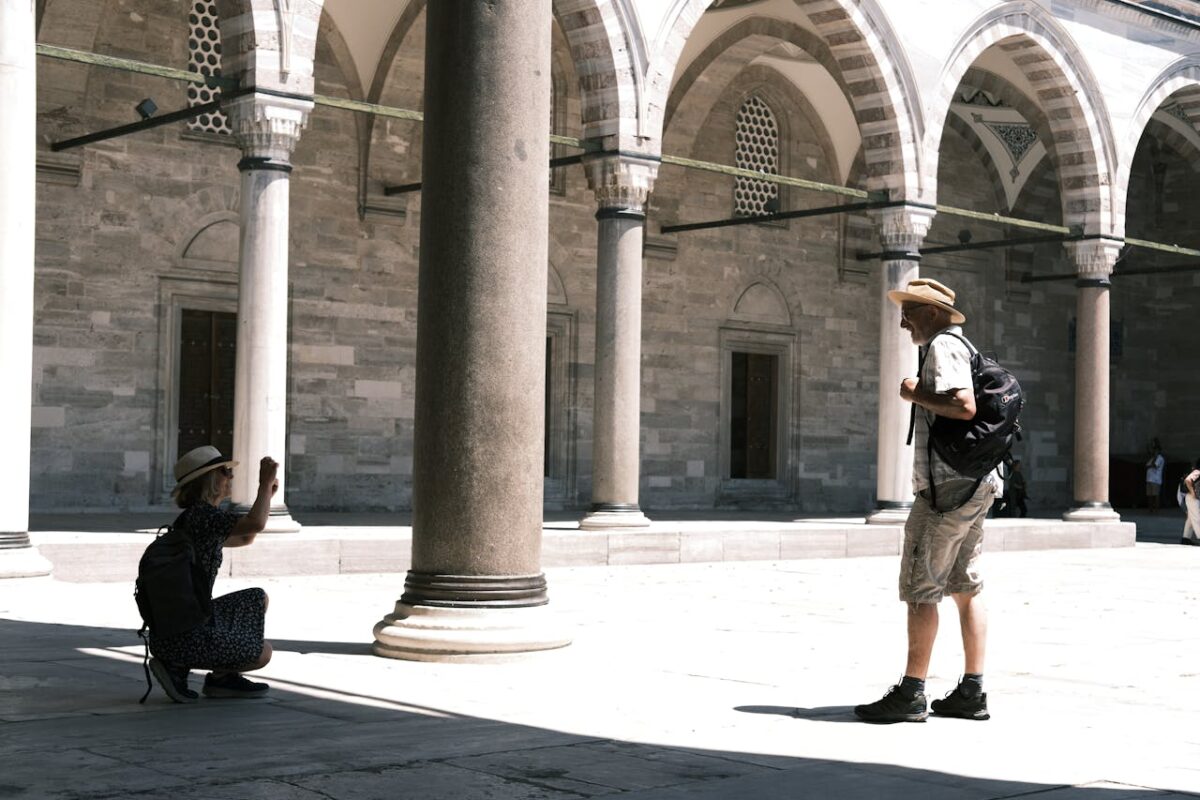 Two tourists take photos in a sunlit courtyard with columns and arches, capturing local architecture.