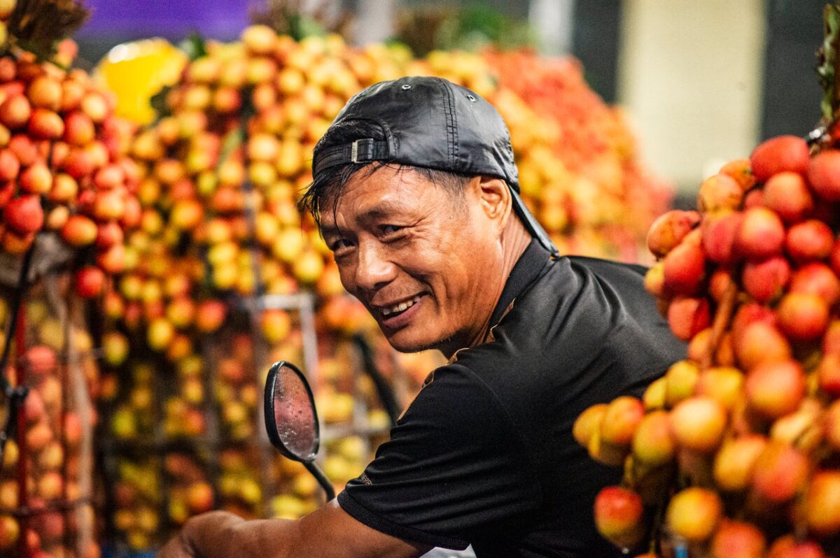 services-01 A joyful vendor at a lychee market in Bac Giang, Vietnam, surrounded by fresh fruit.